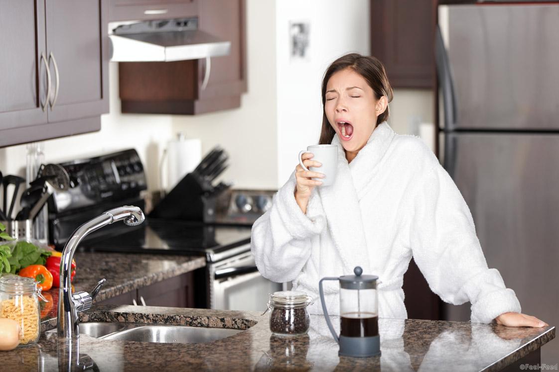 Yawning coffee woman in morning standing by kitchen table at home. Sleepy tired multiracial female model in bathrobe drinking coffee from french press.