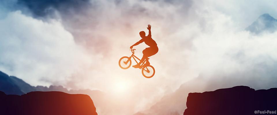 Man jumping on bmx bike over precipice in mountains at sunset. Raising hand showing hello gesture. Extreme sport, risk, cycling.